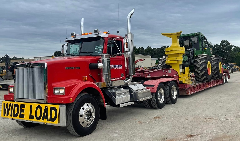 Red semi-truck hauling a green farm tractor. There is a yellow warning sign on the front of the truck with black text that says Wide Load. The sky is cloudy.