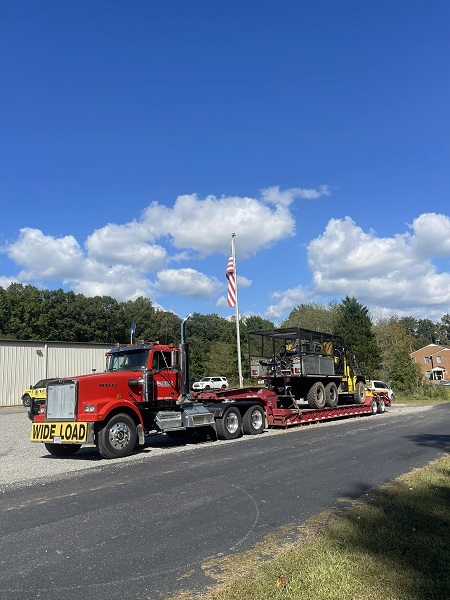 Red tractor trailer rated for wide loads pulling out of a driveway. It a hauling a large yellow vehicle. There is an American flag at the driveway entrance